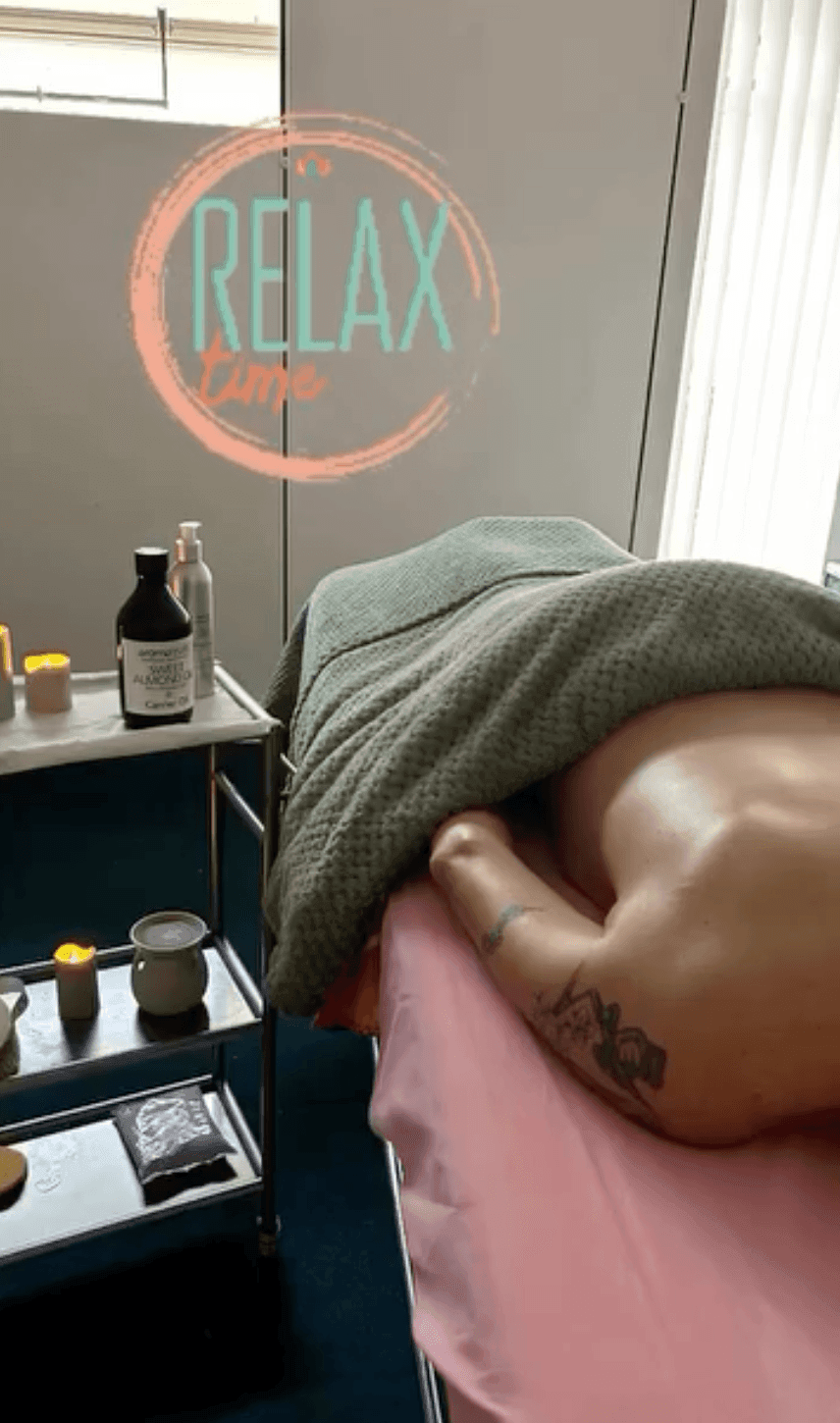 Person lying on massage table with candles and skincare products nearby, 'Relax' text on wall.